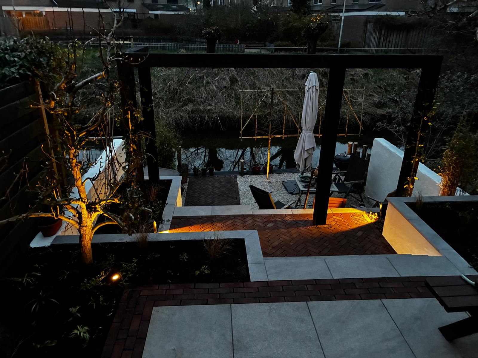 Verlicht verdiept terras met klinkers, een zwarte pergola en verhoogde borders, bij avond