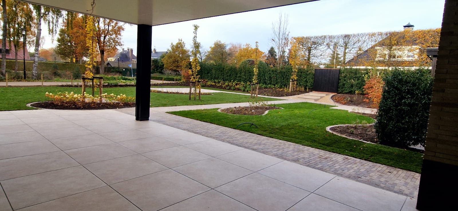 Uitzicht vanonder een overkapping over een groot tegelterras naar een ruime gazontuin met borders en jonge bomen